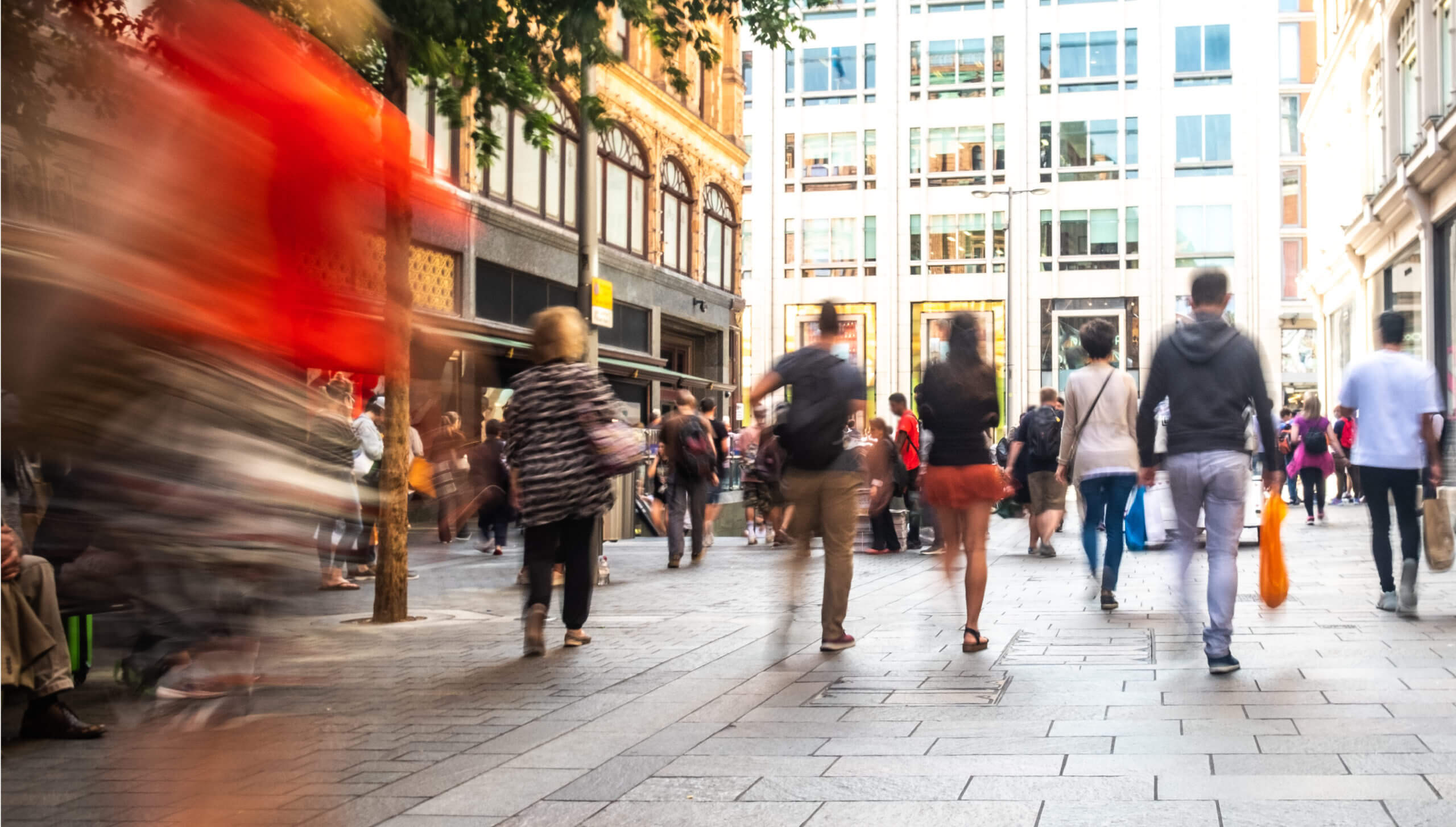 Various people walking in a street with shops either side, some carrying shopping bags. Blue effect on some of the people shown.