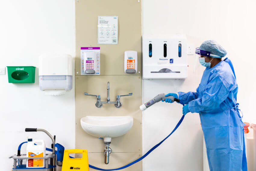 Mitie employee in blue PPE spraying a hospital sink with cleaning fluid