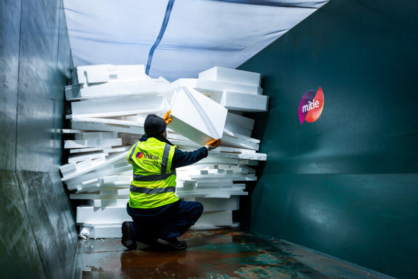 A man in a Mitie hi-vis vest sorting waste polystyrene in a dark green skip