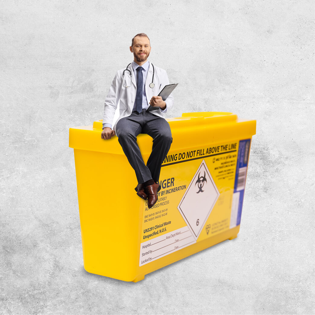 Man wearing a white lab coat and stethoscope sitting on top of an oversized yellow 'sharps' needle bin