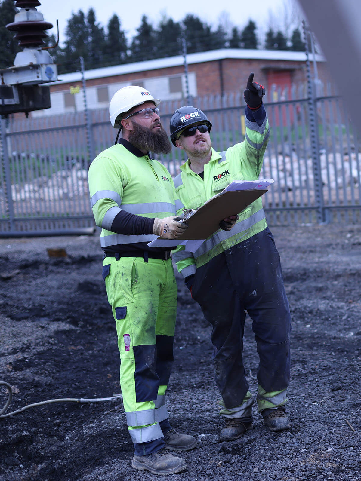 Two male Rock Power Connections employees in high vis clothing, looking at plans and pointing upwards