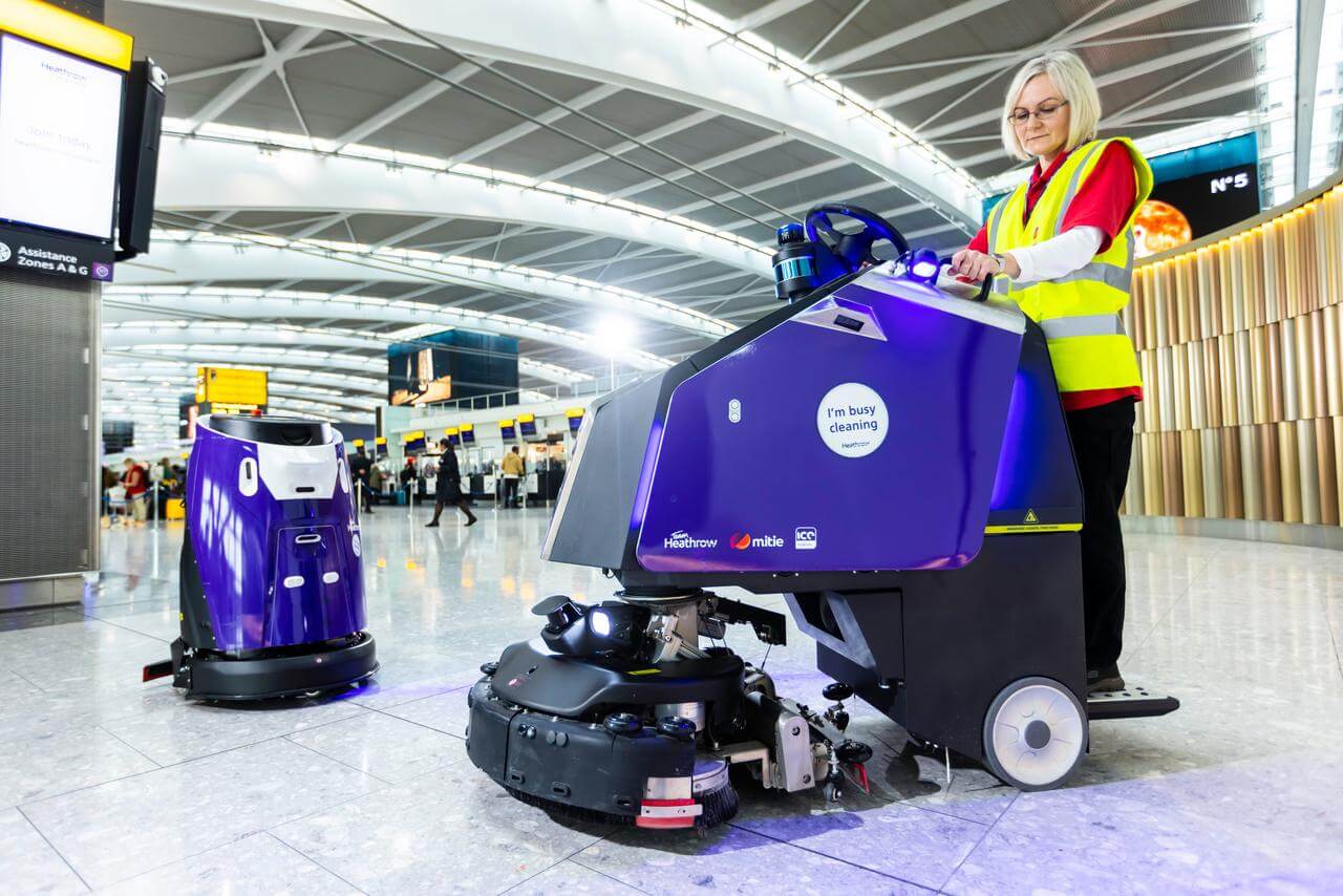 Two purple Mitie cleaning robots, with a Mitie cleaning colleague, in a London Heathrow Airport terminal