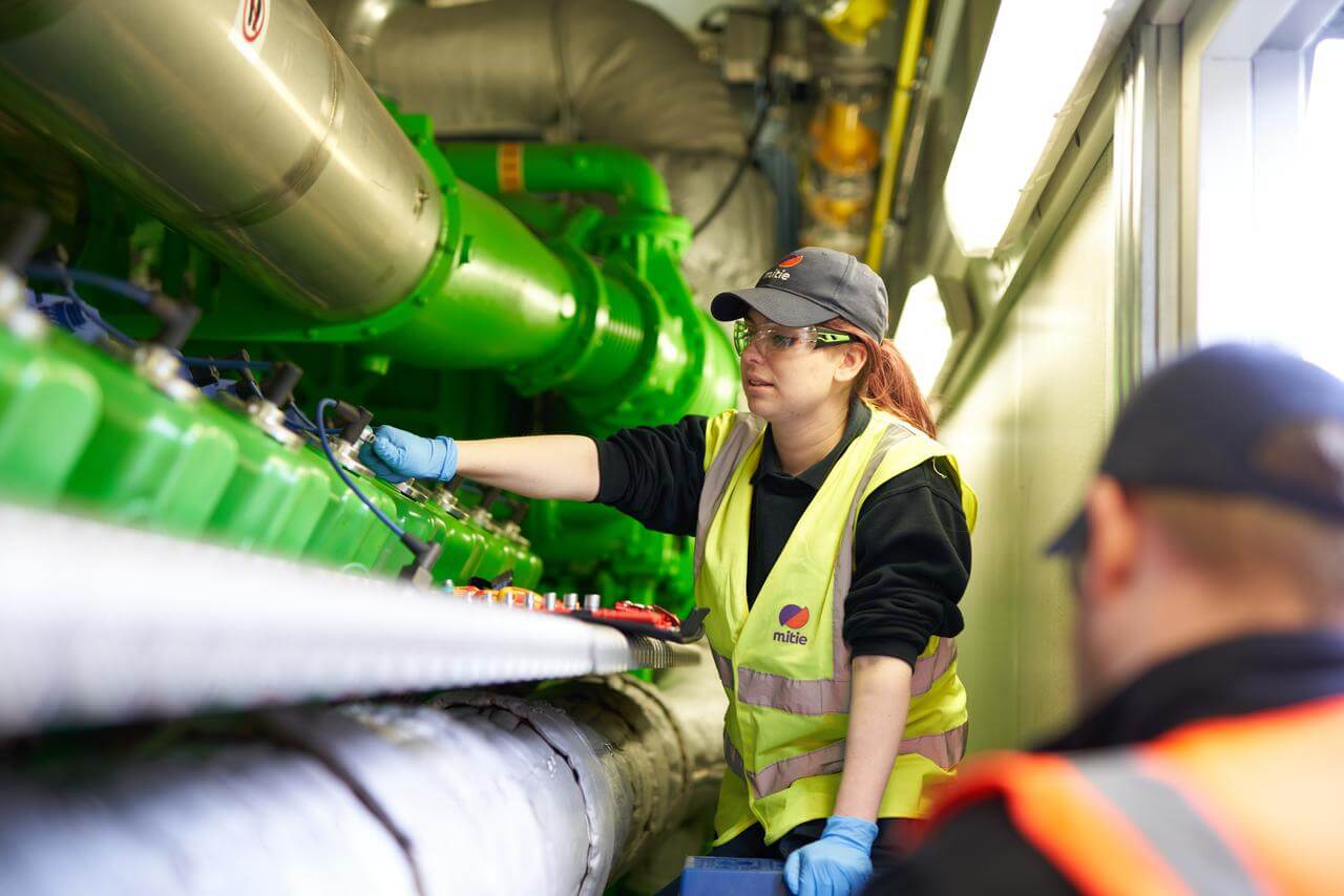 A female Mitie engineer wearing high-vis and safety glasses, working on pipes and equipment