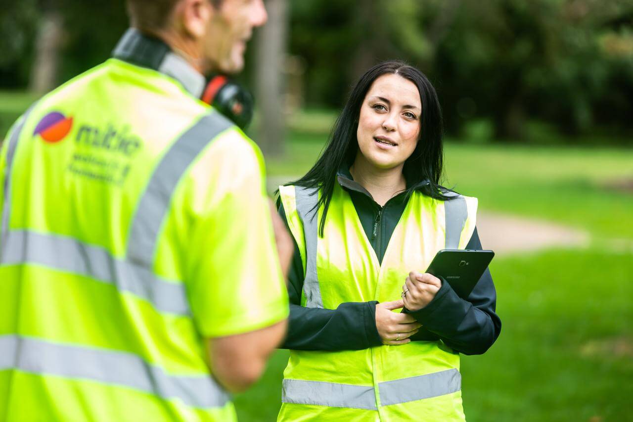 Two Mitie workers in high-vis vests talking in a park