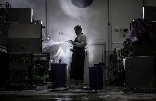Man wearing protective clothing spraying industrial equipment clean, in a dark and steam-filled environment