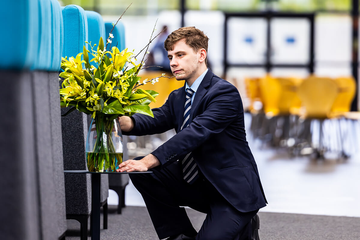 Male front of house employee looking after a vase of yellow flowers on a table in reception area