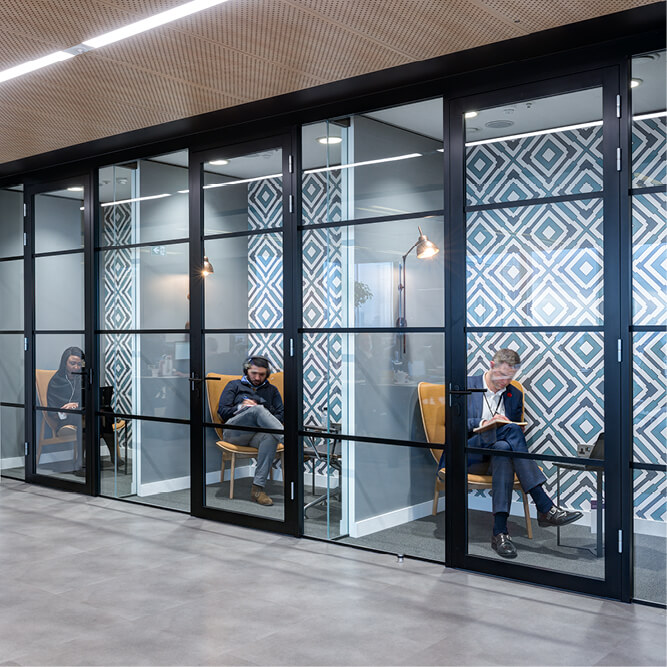 Three individual soundproofed rooms in an office for taking calls, each with a professional person inside sitting on orange chairs