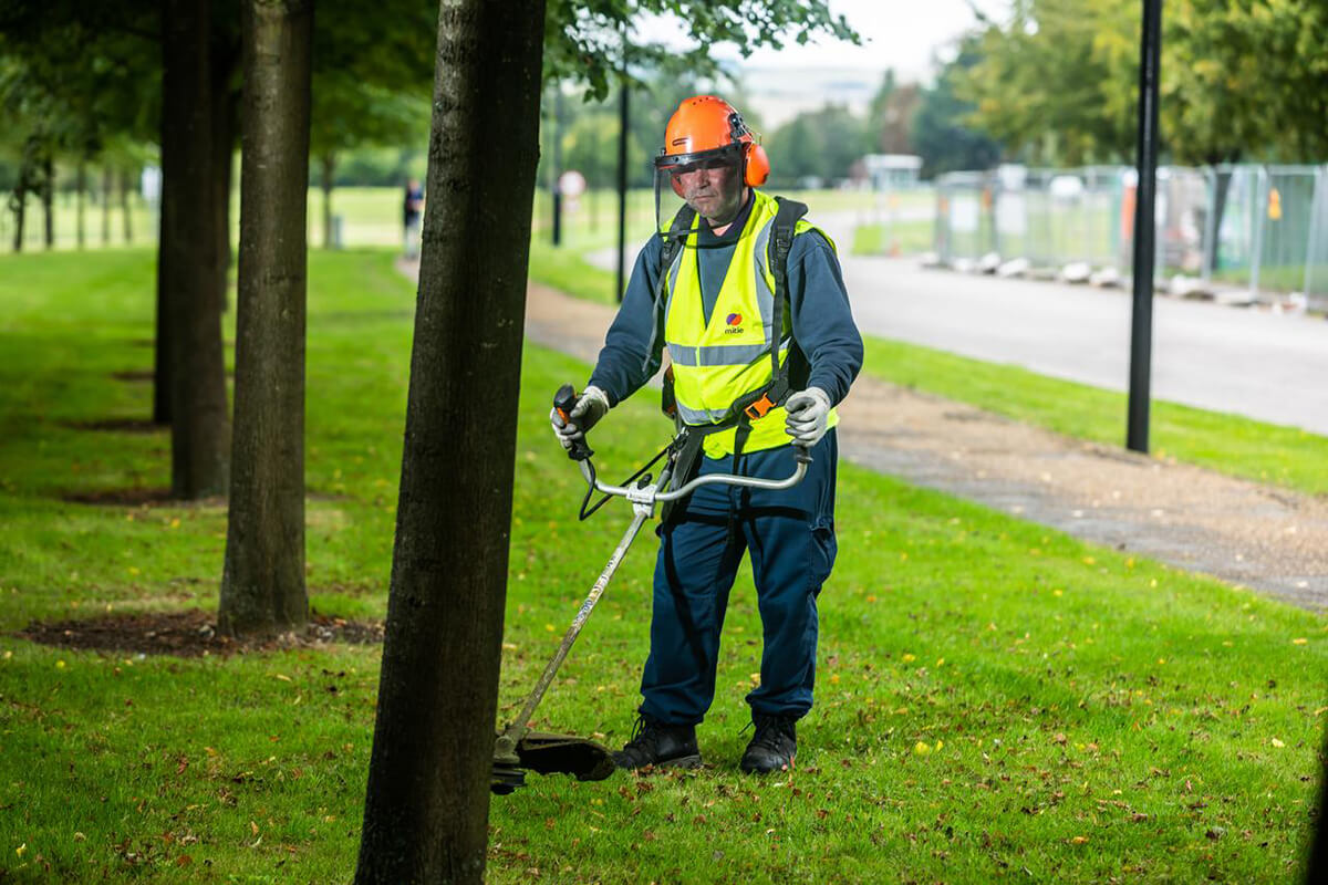 Mitie employee wearing high vis and safety gear using a grass strimmer to conduct ground maintenance