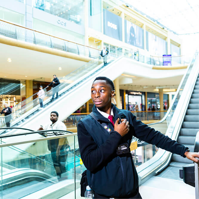 A black male security guard standing at the bottom of an escalator in a shopping centre