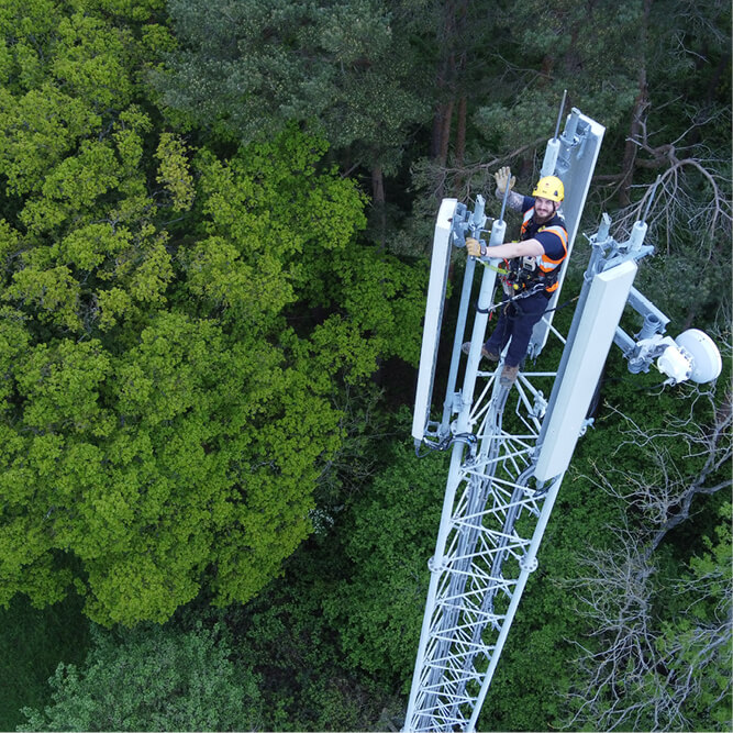 A Mitie engineer wearing safety gear, waving from the top of a telecoms mast