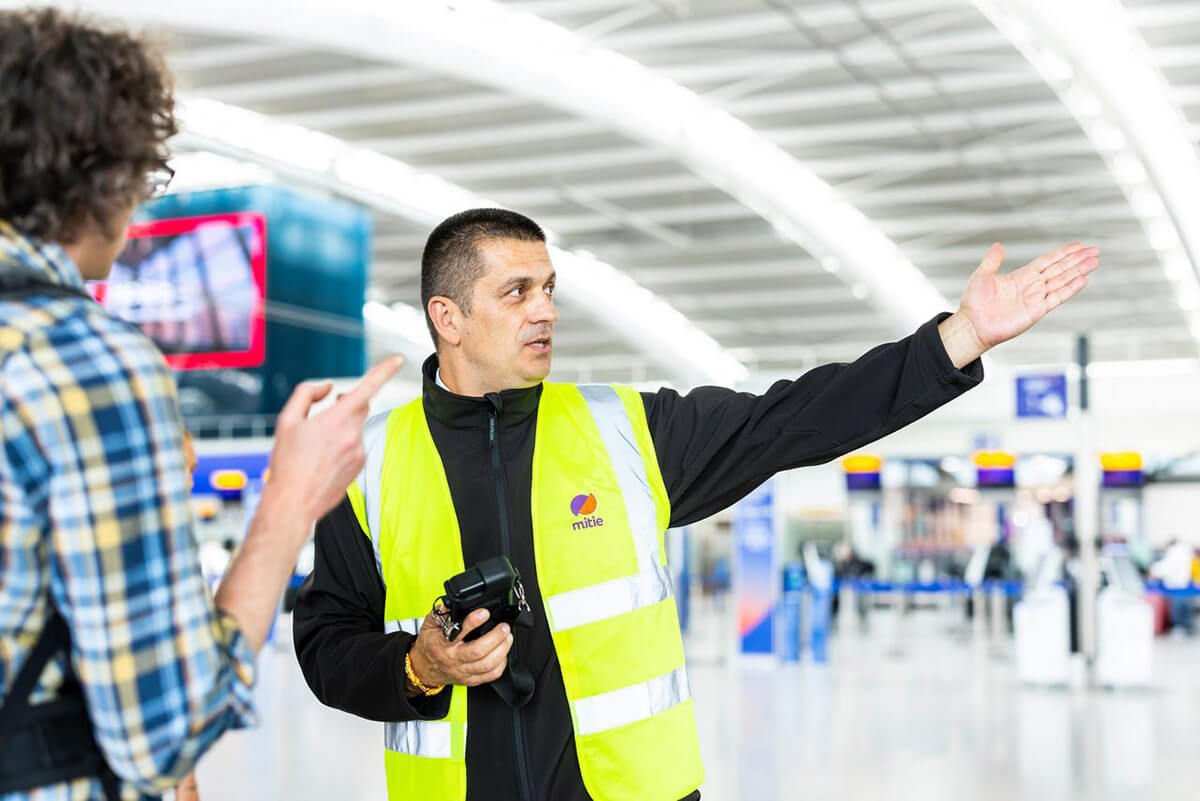 Male security guard at Heathrow Airport, wearing a Mitie branded high vis, pointing to the right and directing a customer