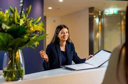 Smiling woman behind a reception desk, offering a sign in book. A vase of yellow flowers are on the desk in the foreground