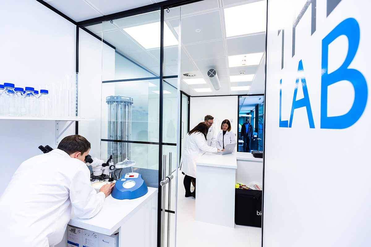 A group of people wearing white lab coats, one looking through a microscope, in a white-walled set of rooms called The Lab in the Mitie Cleaning and Hygiene Centre of Excellence