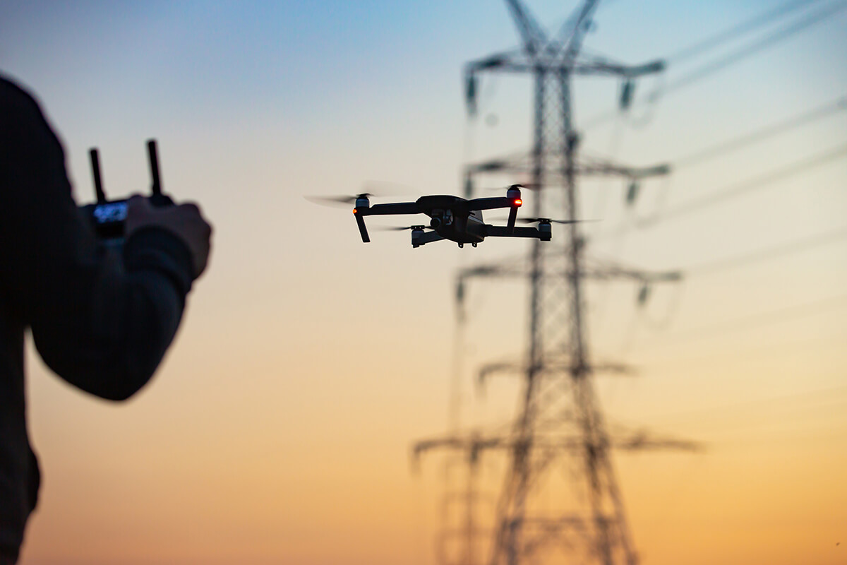A drone flying in front of an electricity mast at sunset, with part of the pilot using the remote in the foreground