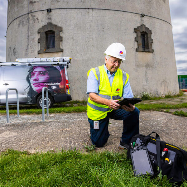 Mitie engineer kneeling on the ground next to a bag of tools, outside a tower in Essex County Council, while looking at an electronic tablet