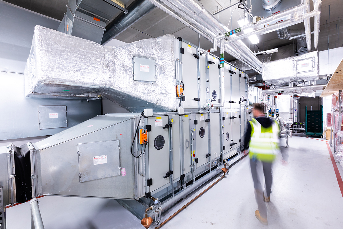 Mitie male employee, blurred and wearing a hi vis vest, walking in a room containing large manufacturing equipment