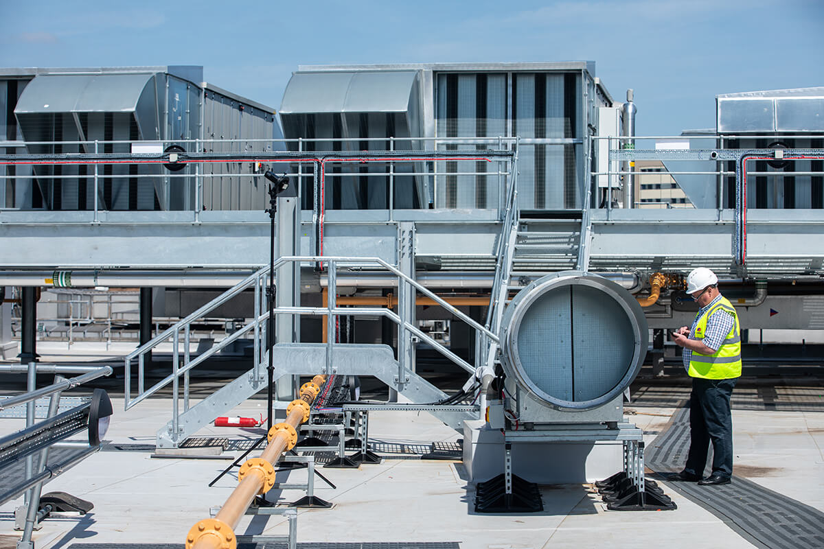 Mitie male employee, wearing a white hard hard and Mitie hi vis vest, inspecting large equipment on a roof