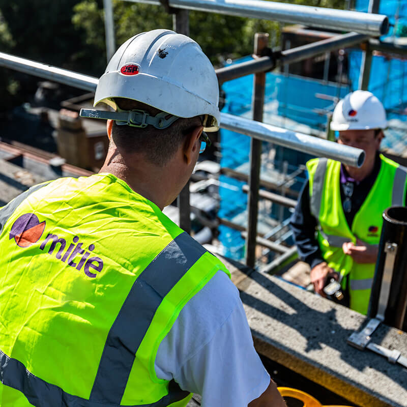 Two Mitie workers on scaffolding, wearing high vis vests and hard hats