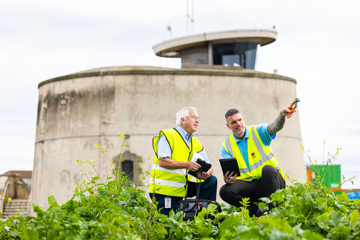 Two male Mitie engineers wearing high vis vests, crouching in front of Martello Tower in Essex. The man on the right-hand side is pointing off-screen with a tool in his hand