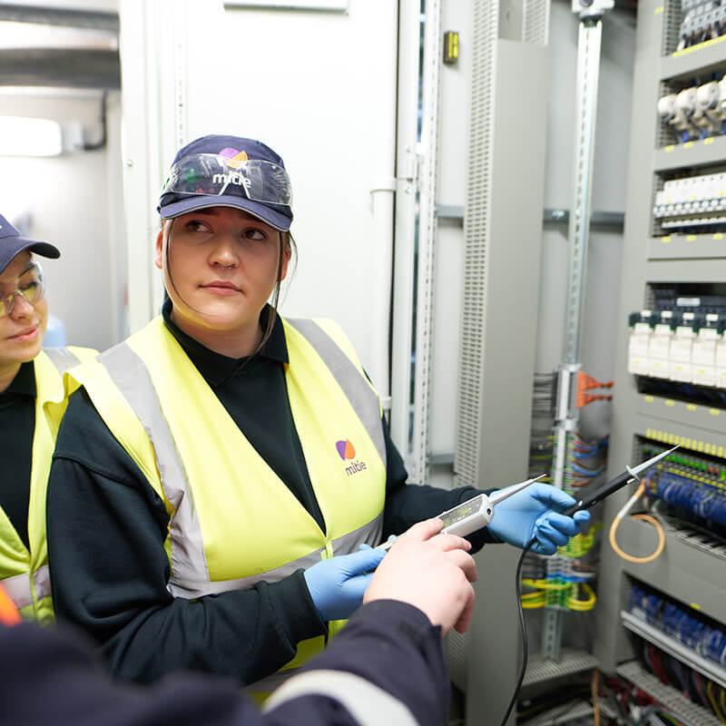 Two female engineer apprentices, wearing Mitie high vis vests and hats, in front of a wall of electrical wires and fuses
