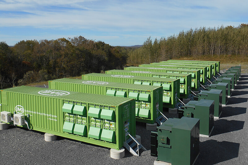 A line of large green corrugated metal containers, used for energy battery storage. They are on an asphalt surface, surrounded by trees and with a blue sky above