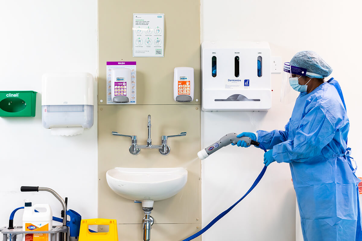 A woman dressed in blue PPE is using a specialist hose to spray a white sink in a clinical setting