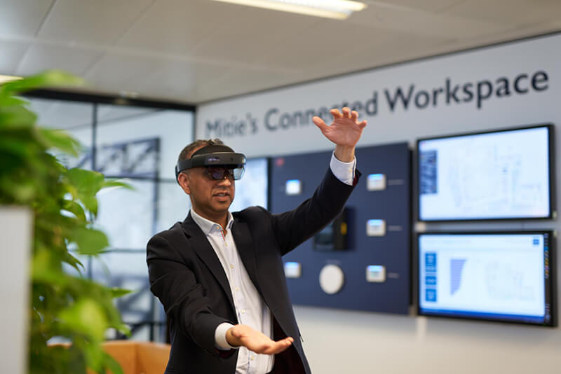 Man, wearing a black suit and white shirt, with a VR headset on his head and waving his arms. 'Mitie's Connected Workspace' is printed on the wall behind him