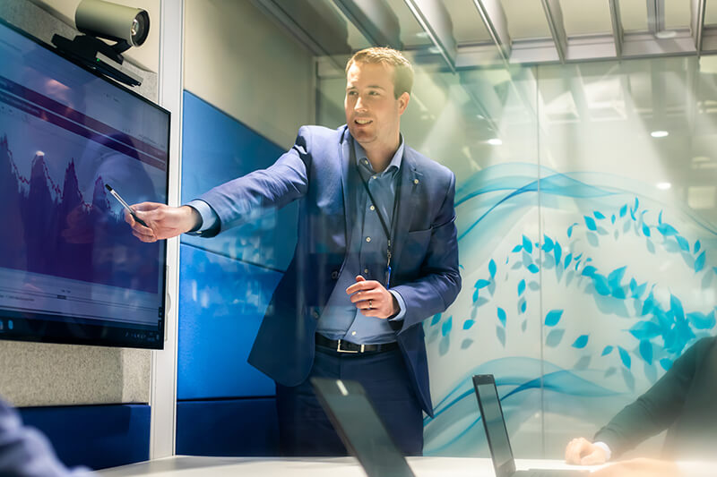 Man wearing a blue suit giving a presentation, pointing at some interesting data on the screen behind him