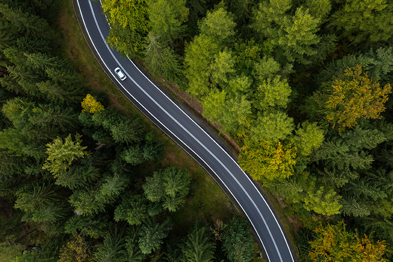 Bird's eye view of a car driving on a bendy road through a surrounding green forest of trees