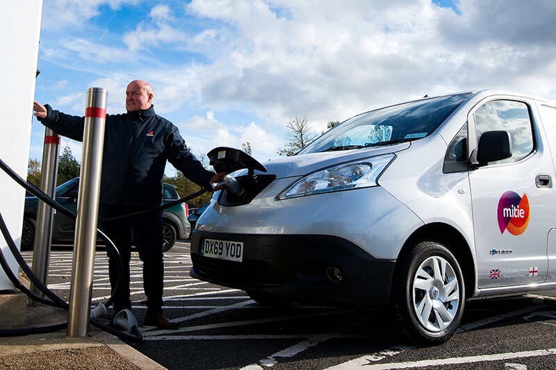 A Mitie branded electric van being charged, with a Mitie employee operating the charge point system at the front of the van