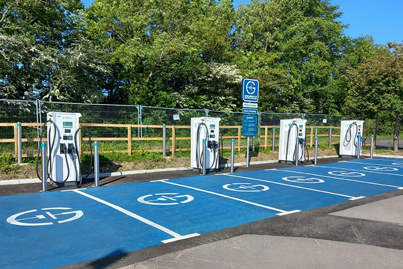 A line of four charging stations for electric vehicles, white boxes with black cables, with blue parking spaces in front of them. A wooden fence and green trees can be seen in the background