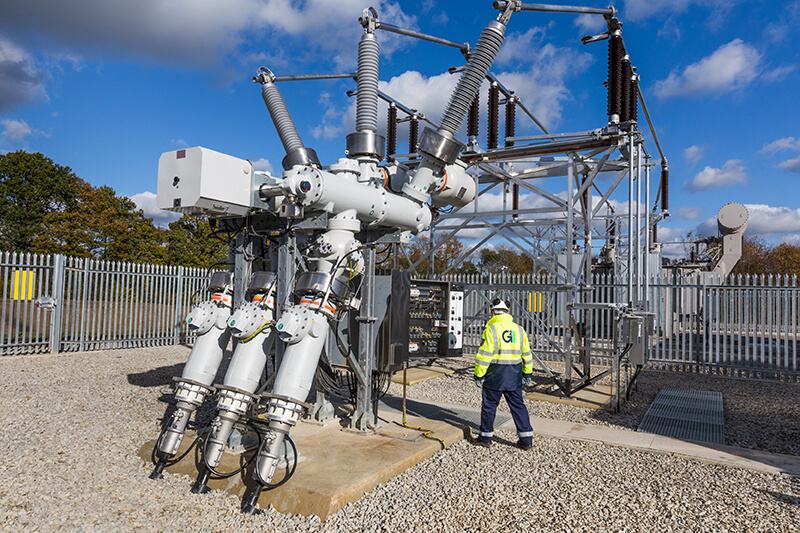 An electrical substation with wires and coils in a metal structure, with a gravel floor and a metal surrounding fence. A G2 Energy worker in high-vis and hard hat is in the middle