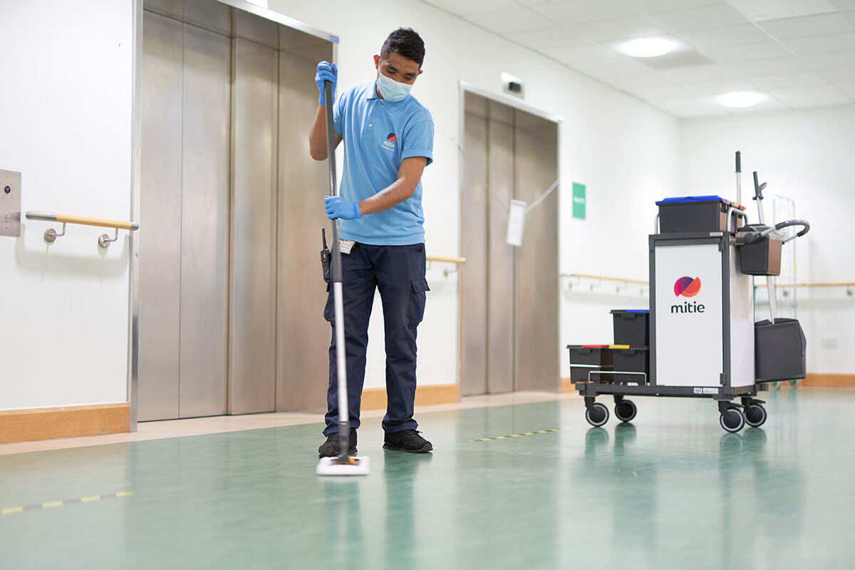 A Mitie employee using a broom to clean the floor in a hospital corridor, with a Mitie branded cleaning cart in the background