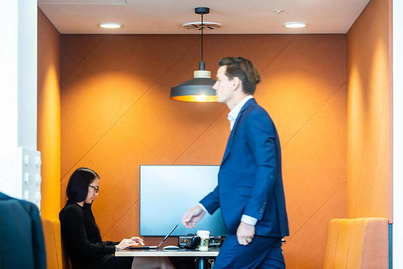 A man in a blue suit walks past an orange seated area in an office, while a woman works on a laptop at the desk