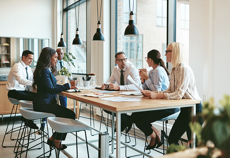 A group of six various people gather round a meeting room table in a modern office environment