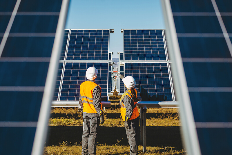 Two engineers in orange hi-ves vests and white hard hats stand in front of a field of solar panels