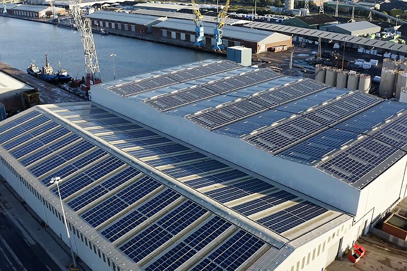 A view of a warehouse roof from a high elevation, with blue solar panels covering the roof. The warehouse is in an industrial area next to a river