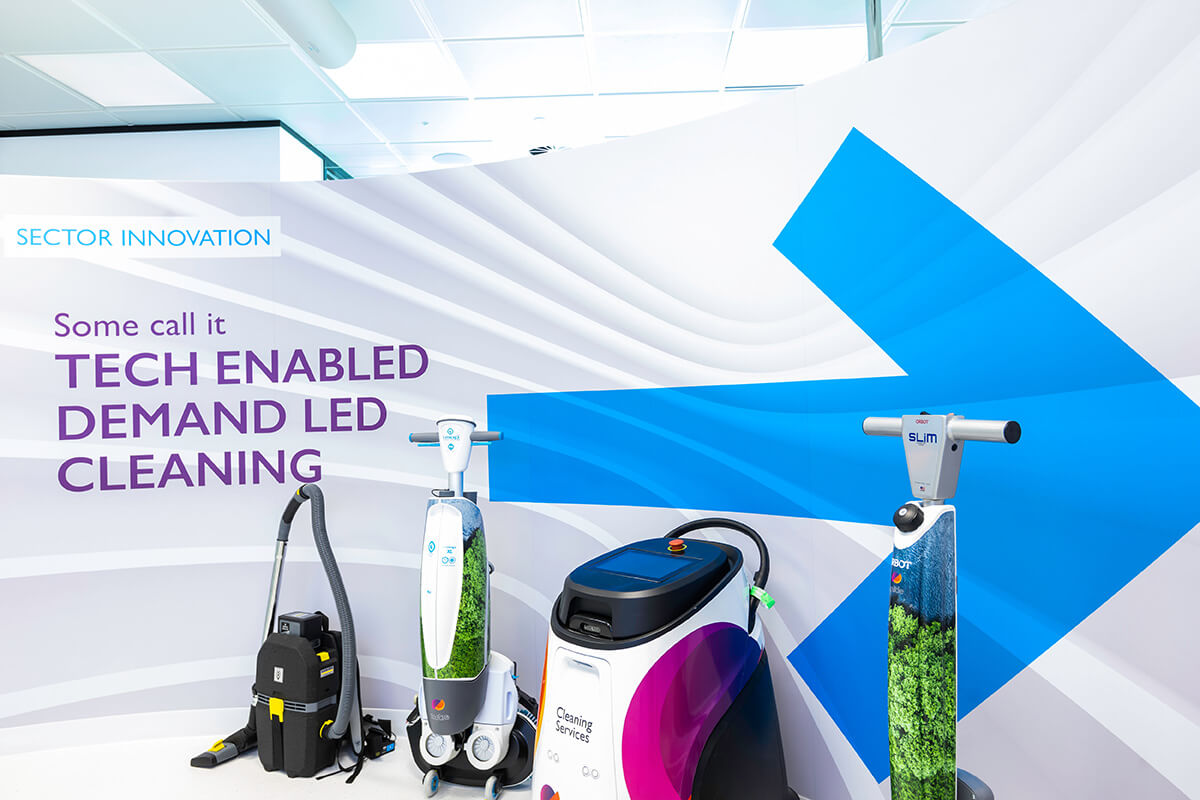 Four different cleaning machines at Mitie's Cleaning & Hygiene Centre of Excellence, in front of wording of the wall behind 'Some call it tech enabled demand led cleaning'
