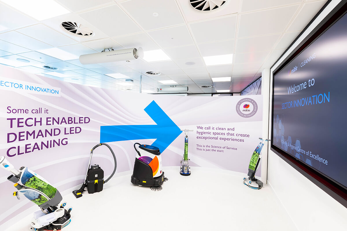 A collection of five cleaning robots and equipment at the Mitie Cleaning & Hygiene Centre of Excellence. The wall behind has text of 'Some call it tech enabled demand led cleaning'
