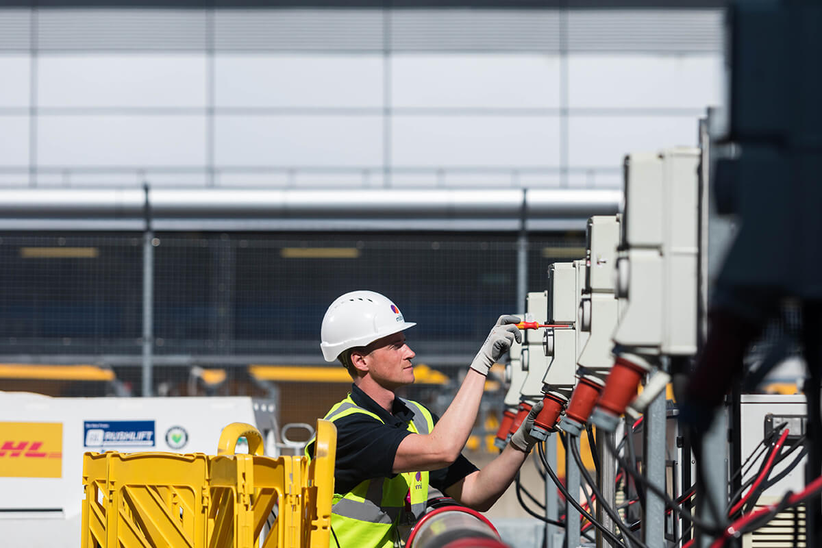 An engineer using a screwdriver to fix an electrical charge point outside of London Heathrow Airport. He is wearing a Mitie-branded hard hat and high vis vest, with yellow safety barriers behind him
