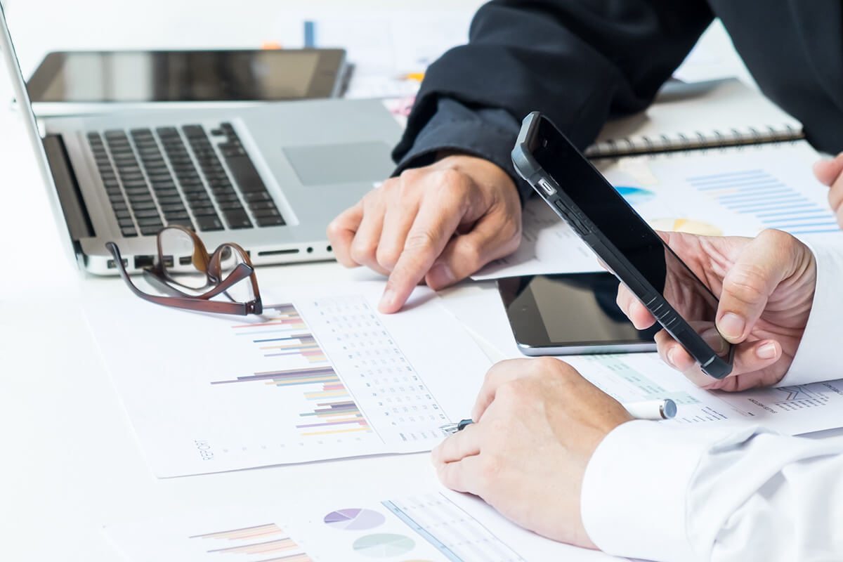 Close up of two people's hands holding smartphones and pointing at charts printed out on paper laying on the desk