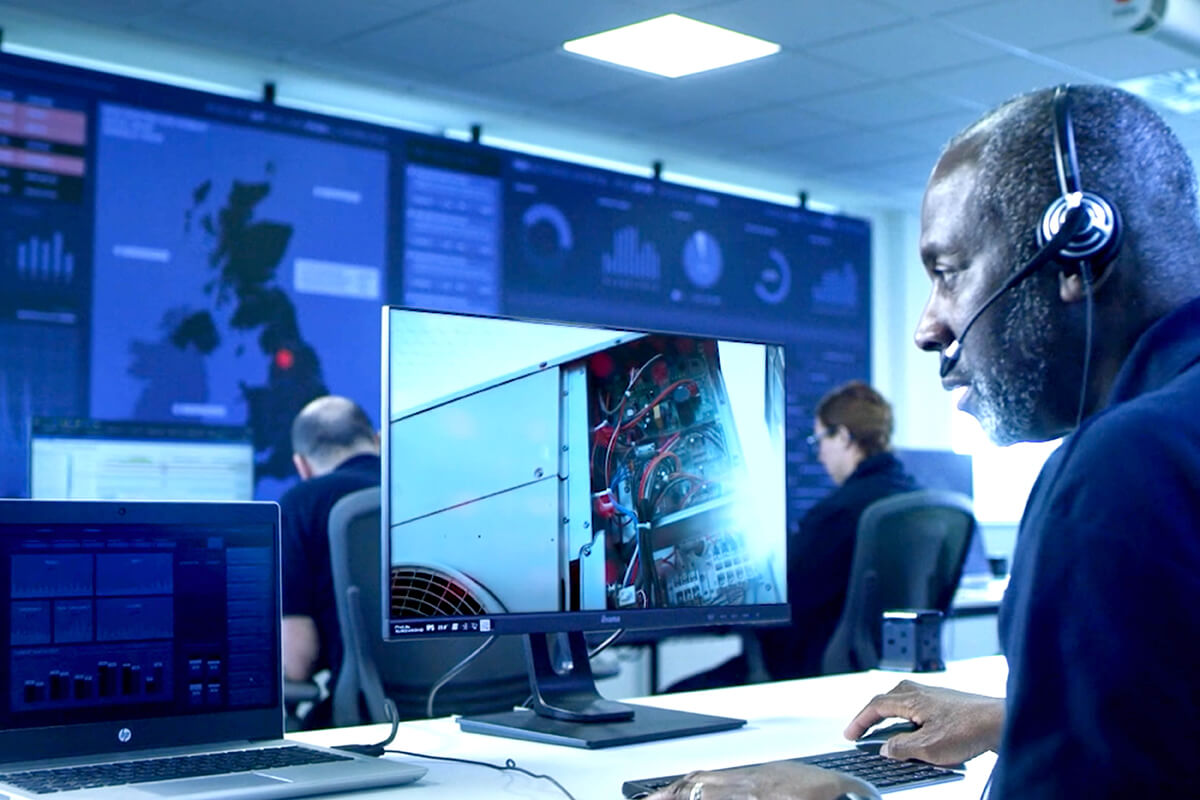 An engineer wearing a headset at a computer terminal, watching maintenance taking place elsewhere. A wall of computer displays and other people at desks are in the background