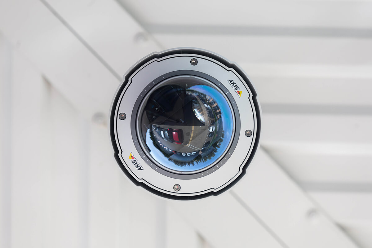 A close-up of a security camera against a white wall. The camera is Axis branded and the reflection of a car park can be seen in the camera
