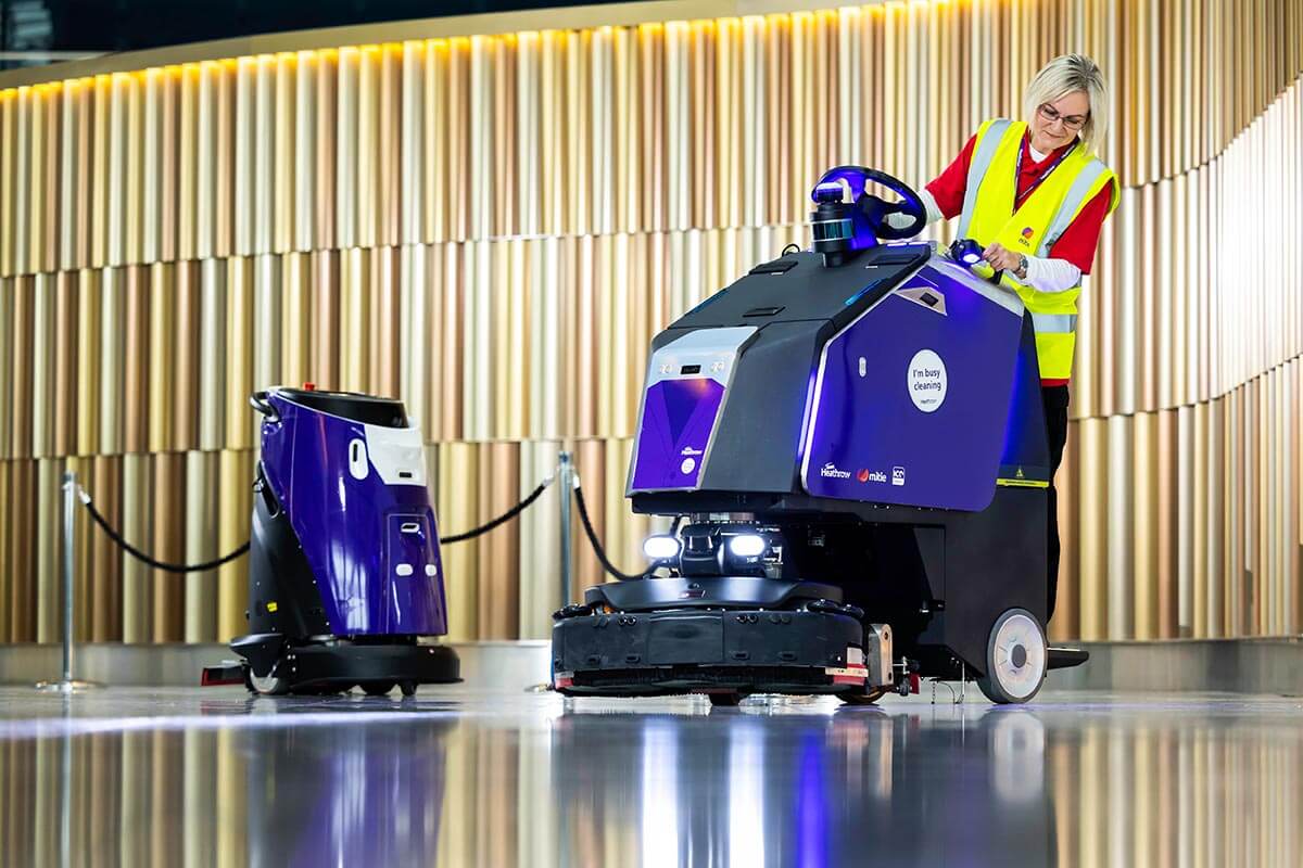 Two purple and black Mitie cleaning robots in London Heathrow Airport, with a female Mitie worker in a high vis vest using the right-hand robot