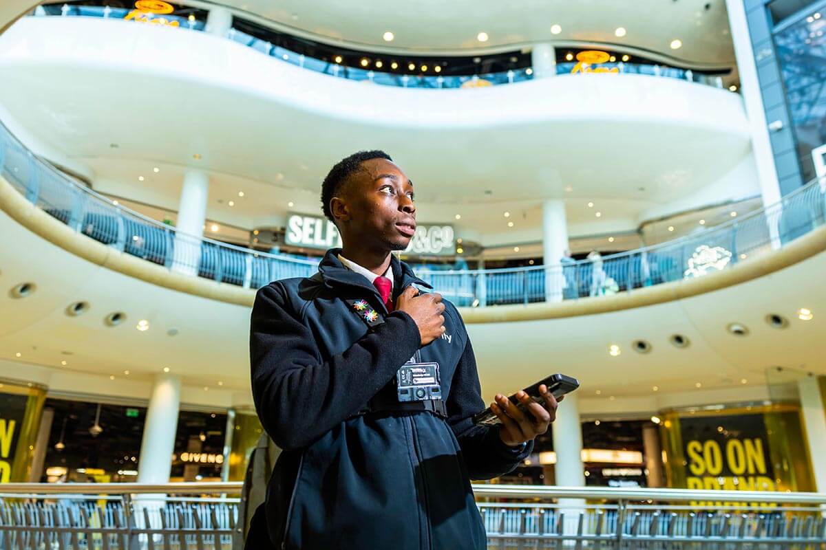 A young black security guard holding his radio, with levels of a shopping centre seen behind him