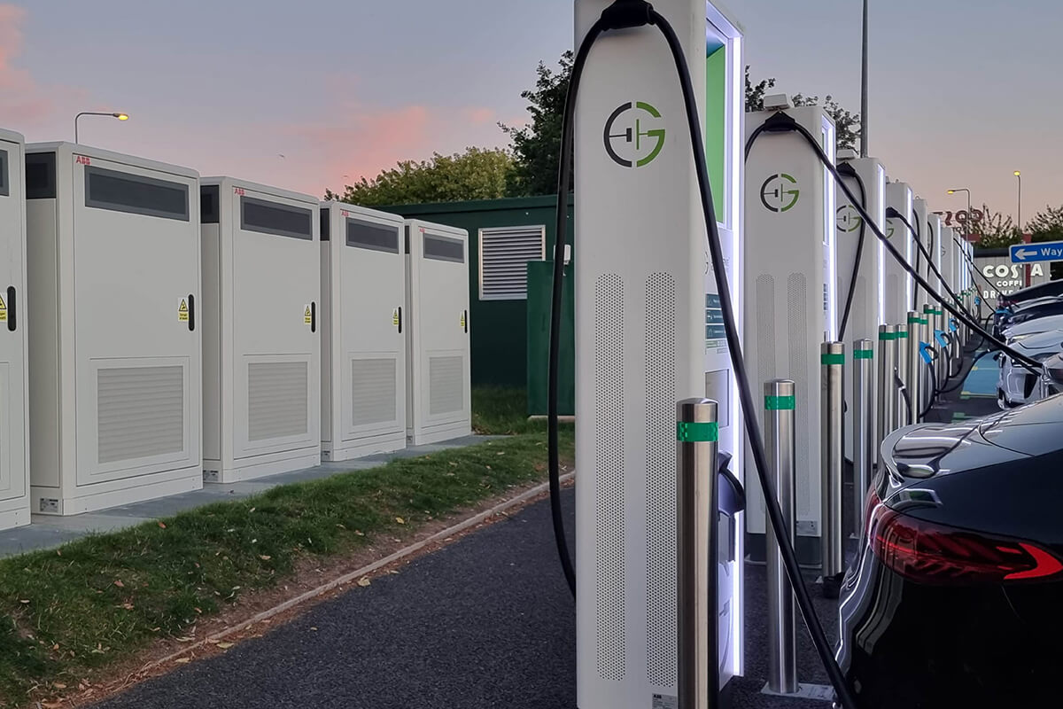 A line of white EV charging points in a car park, with various cars plugged in