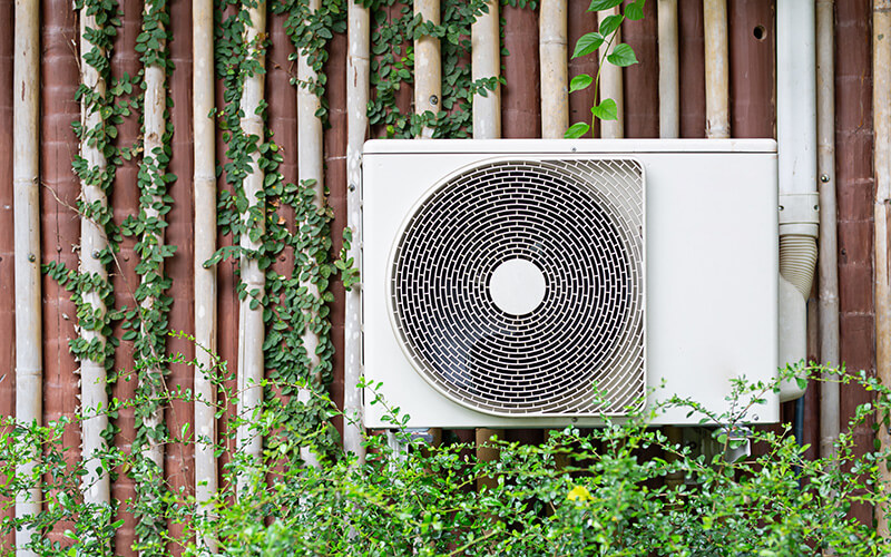 A white air conditioning unit mounted on a wall with greenery surrounding it