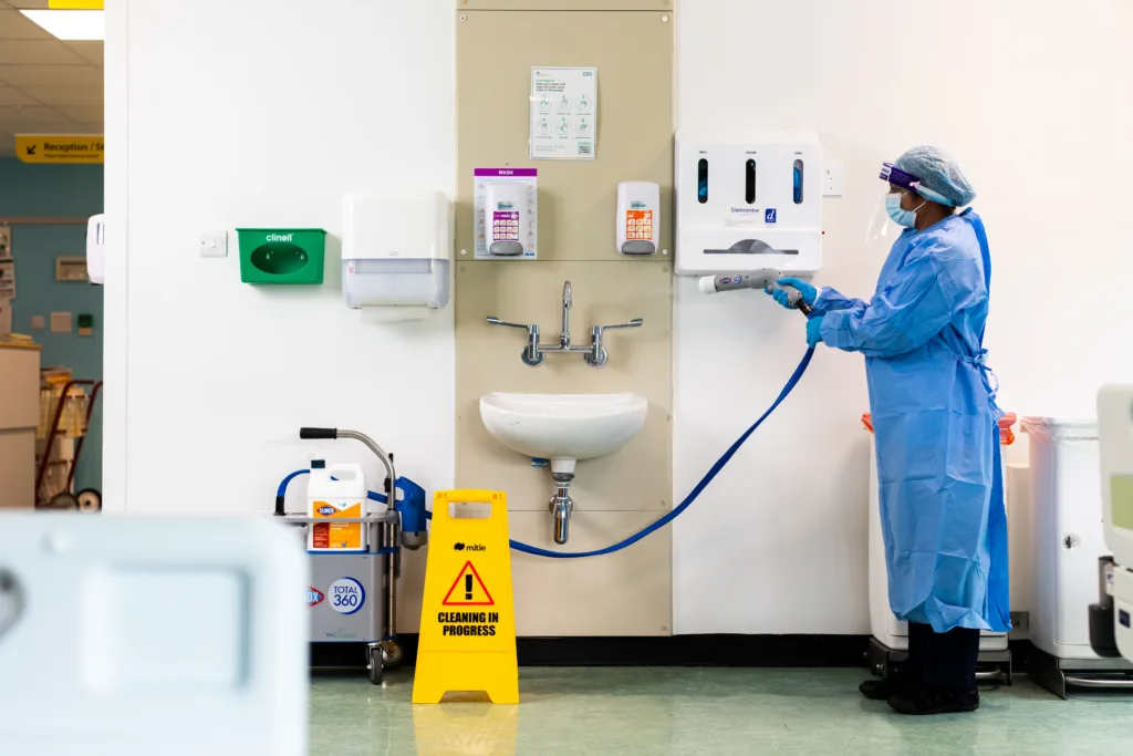 A cleaner in PPE spraying a sink in a clinical setting