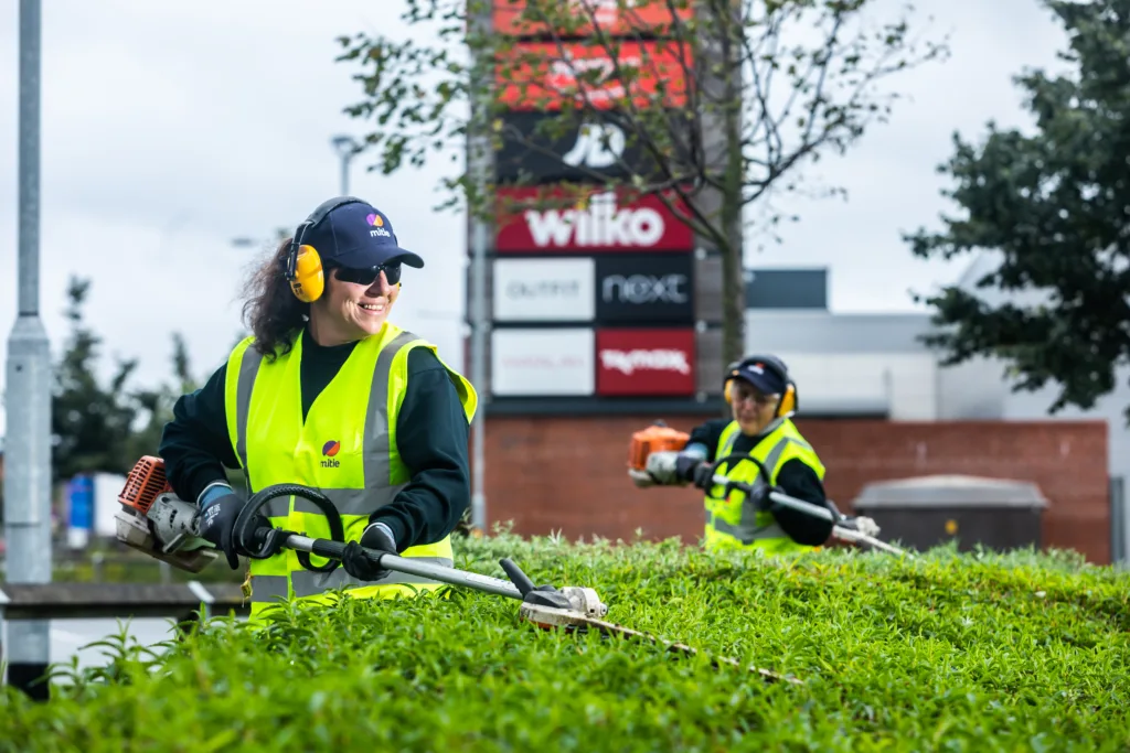 Two women wearing Mitie-branded high vis jackets, hats and safety glasses, strimming the top of a hedge