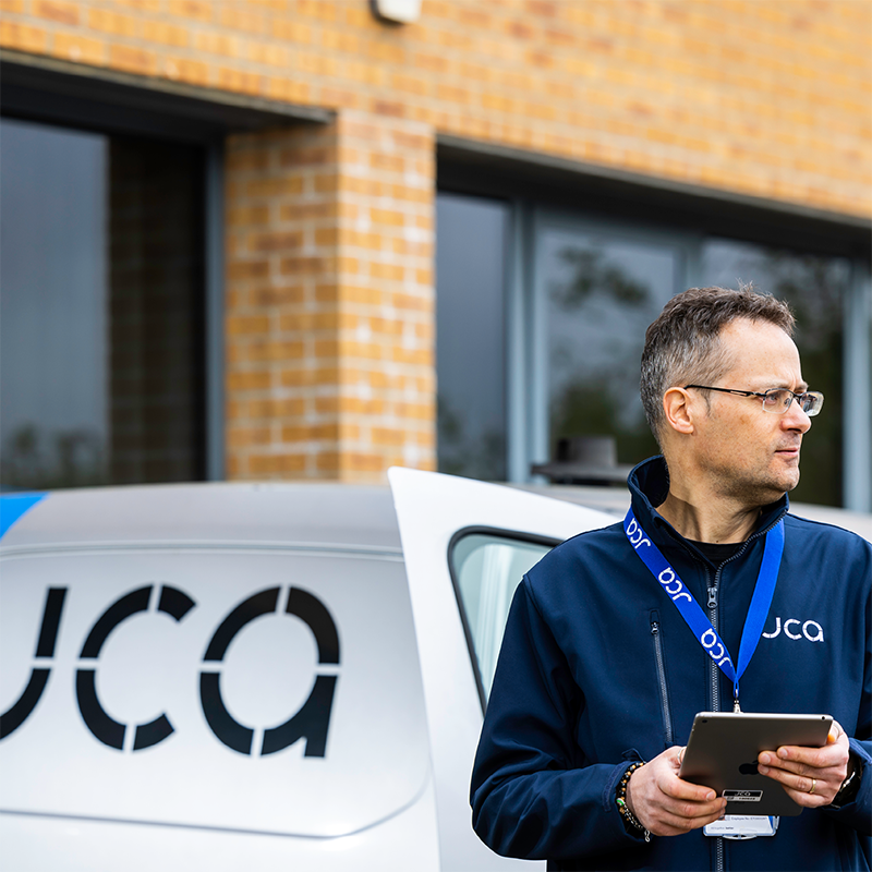 An engineer holding an iPad and wearing a lanyard stands in front of a JCA Engineering branded van